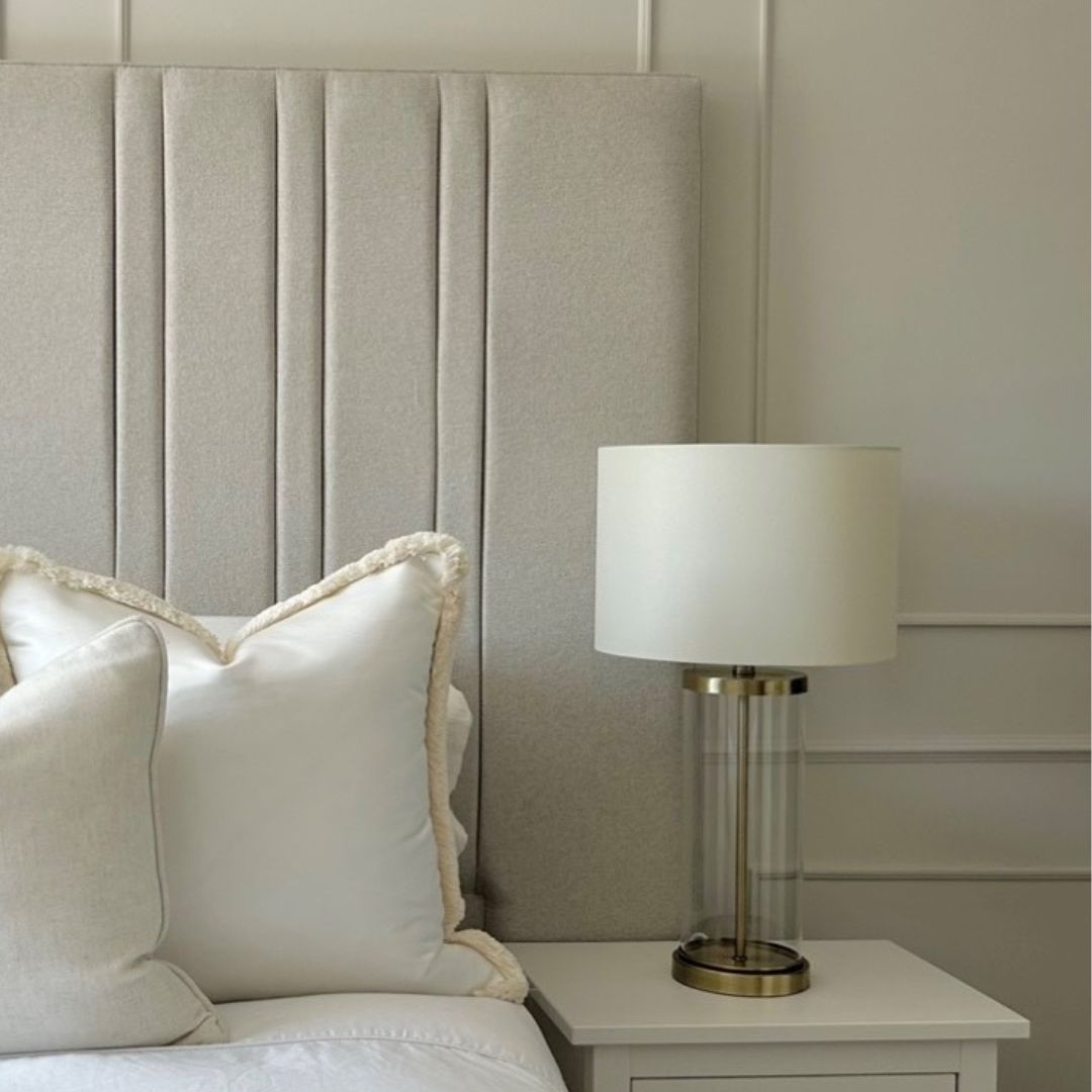 A table lamp with a clear glass base and ivory shade, with a brushed gold effect finish, placed on a bedside table next to a bed with white pillows.